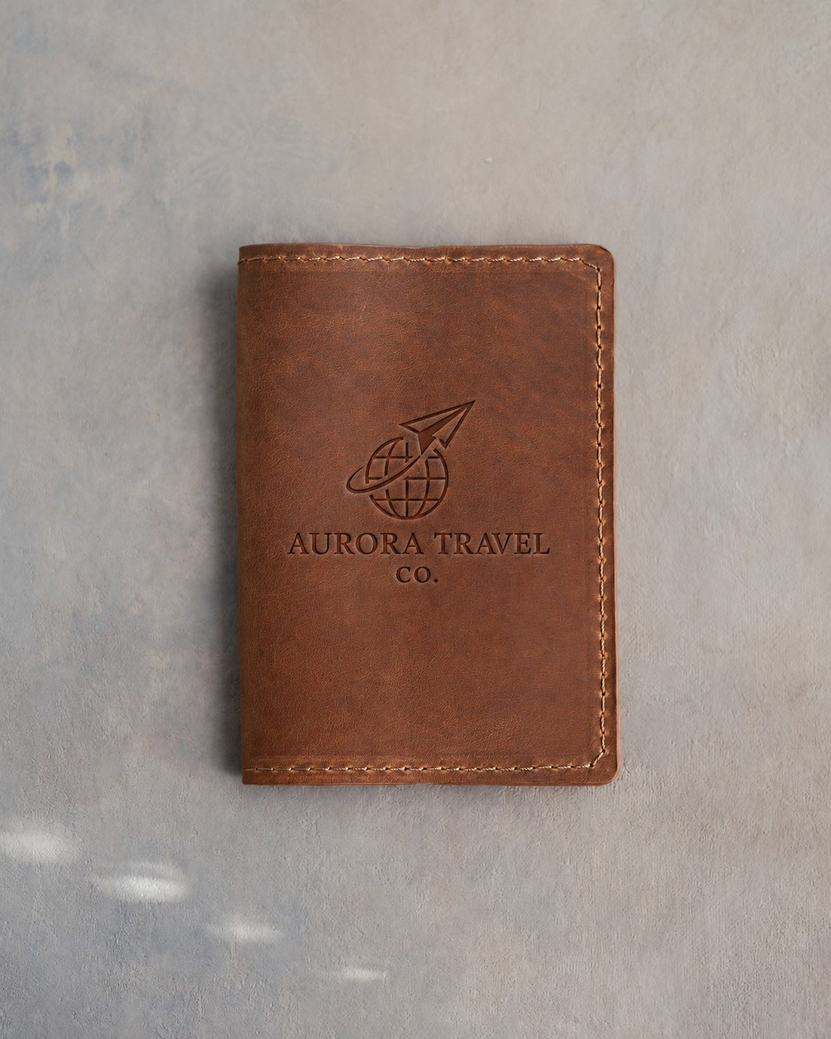 Sicilian Brown leather passport cover with a sample travel agency logo, AURORA TRAVEL CO., pressed into the front, on grey plaster