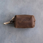 Sicilian Brown leather zipper poop bag holder with a stamped name and a trigger snap clip, on grey plaster
