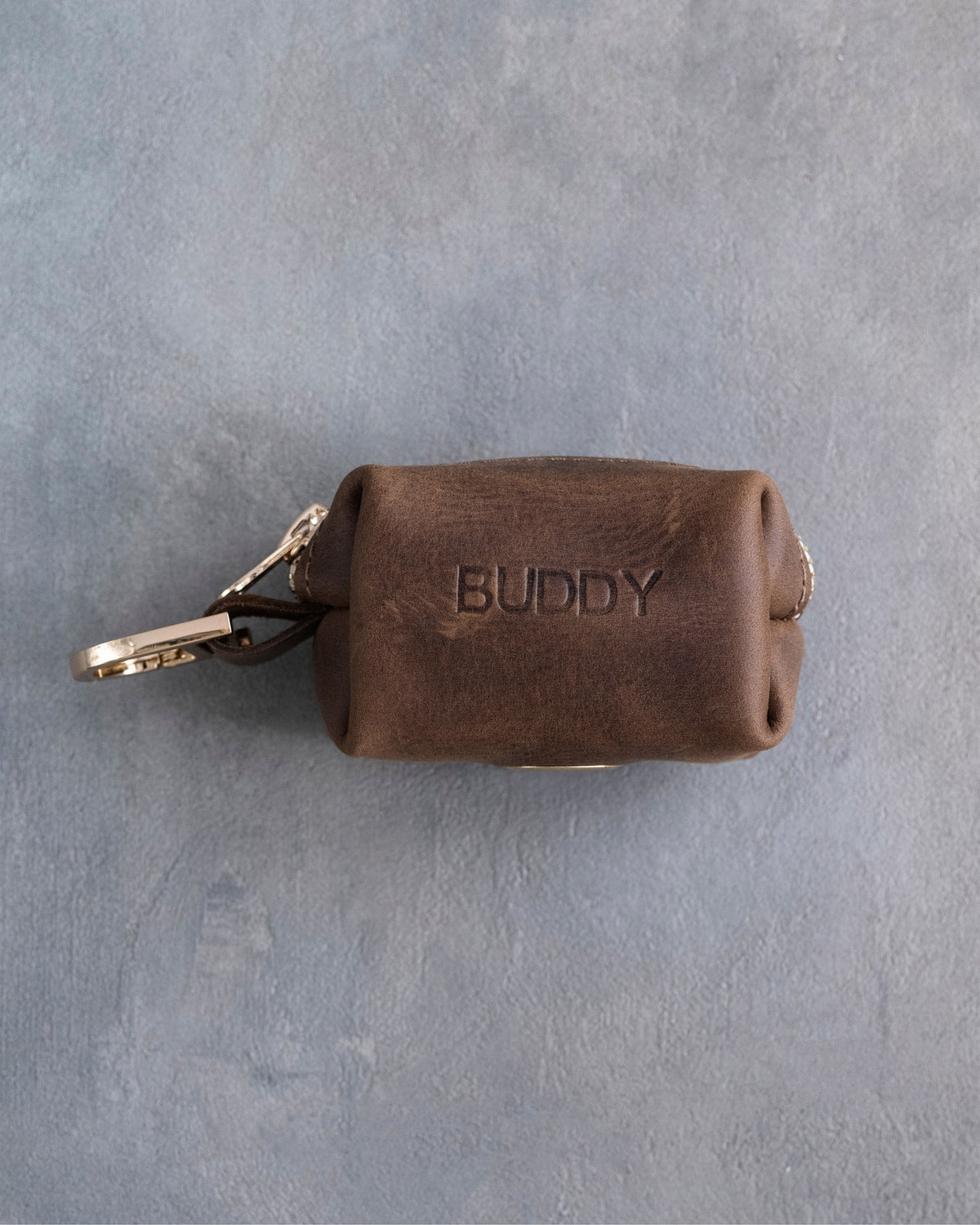 Sicilian Brown leather zipper poop bag holder with a stamped name and a trigger snap clip, on grey plaster