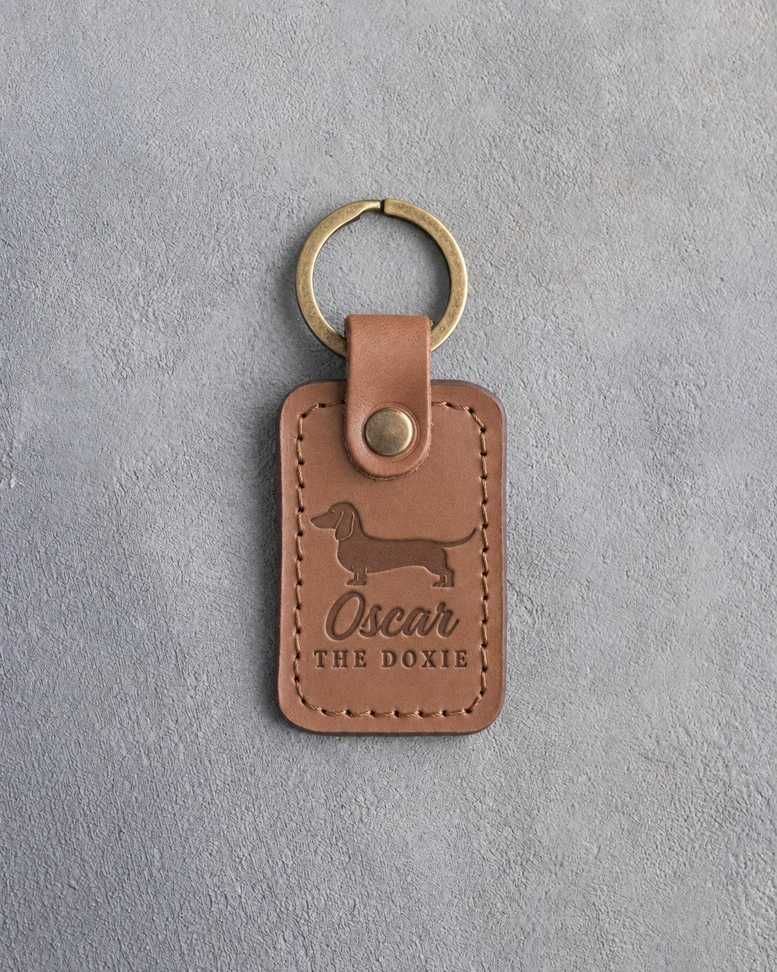 Caramel leather keychain with a dachshund silhouette and the name Oscar above THE DOXIE, debossed, on gray concrete
