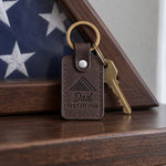 Dark brown Dad Served 1968 keychain on an older man's key ring resting on a wooden mantel