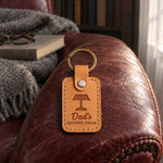 Tan Dad's READING CHAIR keychain on the arm of a worn oxblood leather armchair beside a closed book