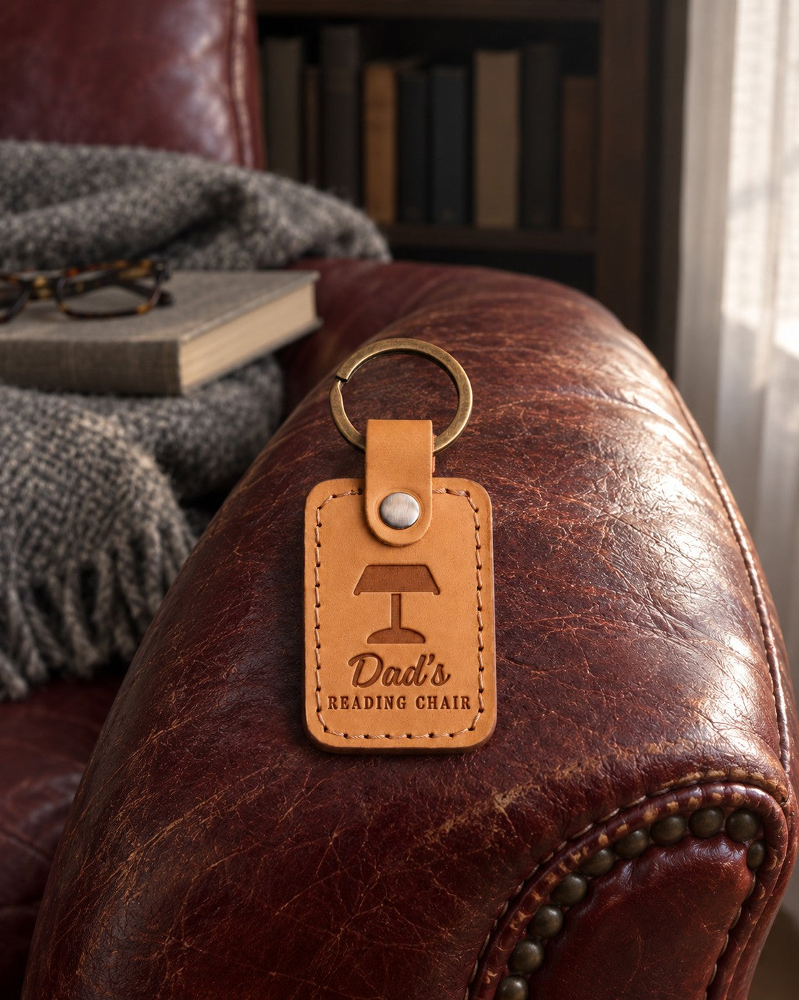 Tan Dad's READING CHAIR keychain on the arm of a worn oxblood leather armchair beside a closed book
