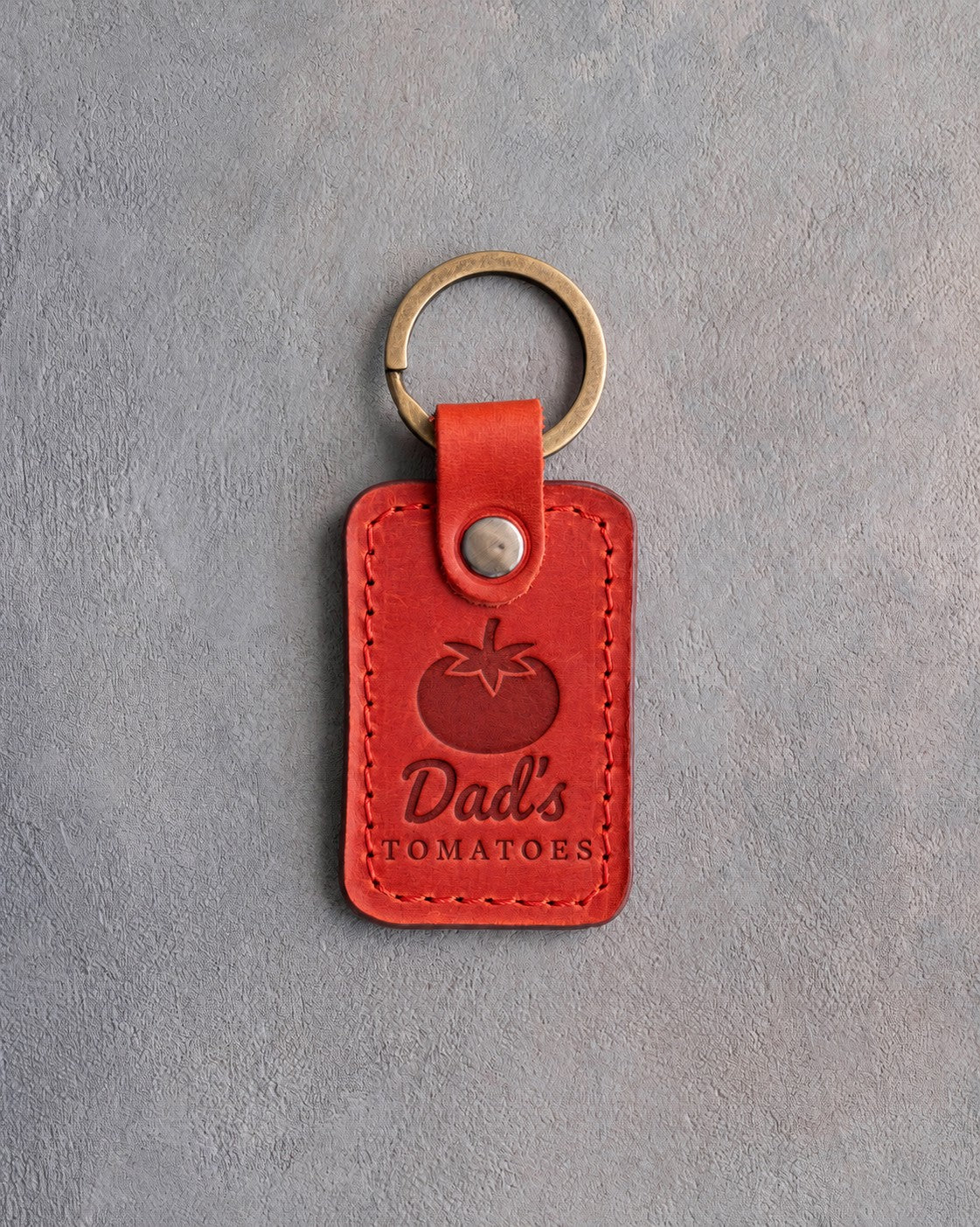 Poppy red leather keychain debossed with a ripe tomato above Dad's TOMATOES, on gray concrete