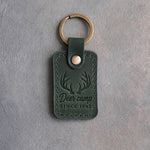 Forest green leather keychain debossed with whitetail antlers above the words Deer camp SINCE 1962, on gray plaster