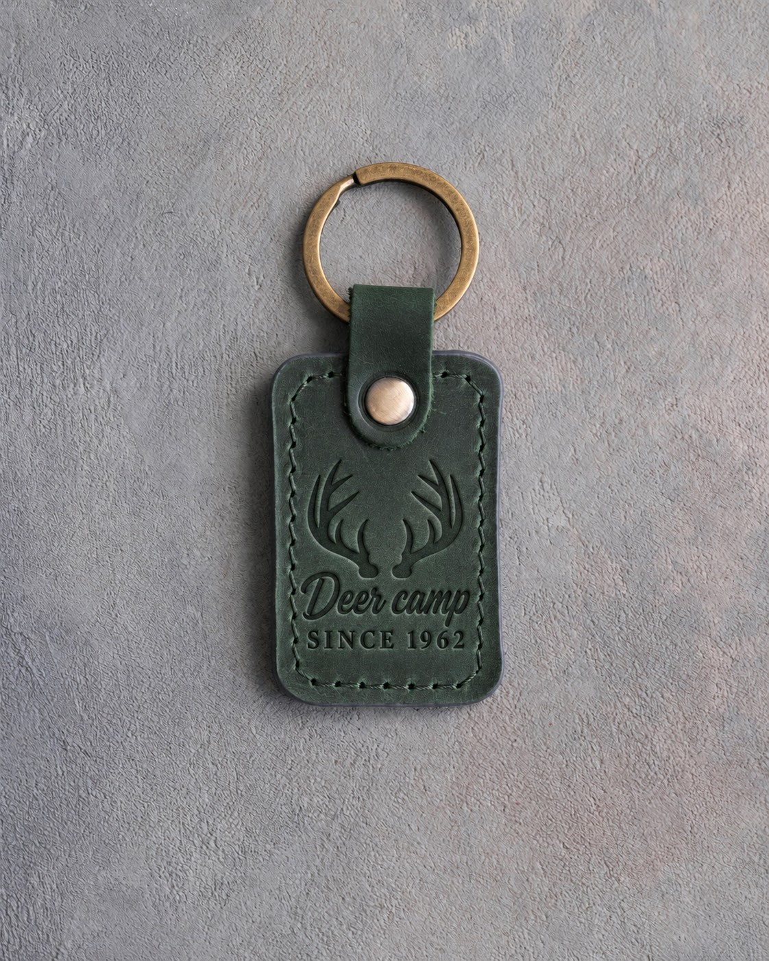 Forest green leather keychain debossed with whitetail antlers above the words Deer camp SINCE 1962, on gray plaster