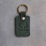 Forest green leather keychain debossed with whitetail antlers above the words Deer camp EVERY NOVEMBER, on gray concrete