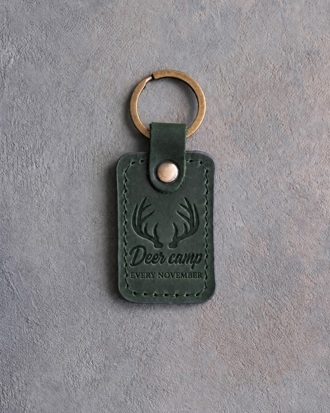 Forest green leather keychain debossed with whitetail antlers above the words Deer camp EVERY NOVEMBER, on gray concrete