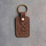 Sicilian brown leather keychain debossed with a crossed trowel and hand fork above Dirt THERAPY, on gray concrete