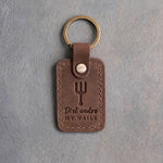 Sicilian brown leather keychain debossed with an upright pitchfork above the words Dirt under MY NAILS, on gray concrete