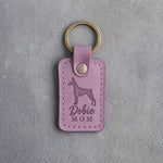 Lilac leather keychain debossed with a Doberman silhouette and the words Dobie MOM, on gray concrete