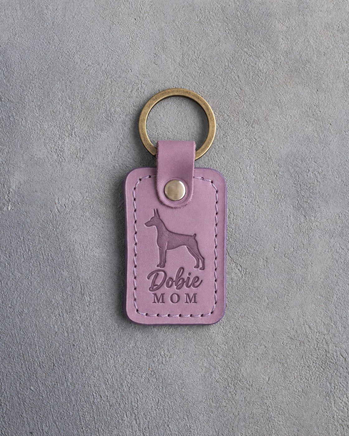 Lilac leather keychain debossed with a Doberman silhouette and the words Dobie MOM, on gray concrete