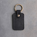 Black leather keychain debossed with a Doberman silhouette above THE DOBIE, on gray concrete