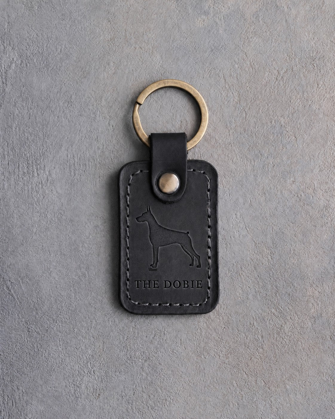 Black leather keychain debossed with a Doberman silhouette above THE DOBIE, on gray concrete