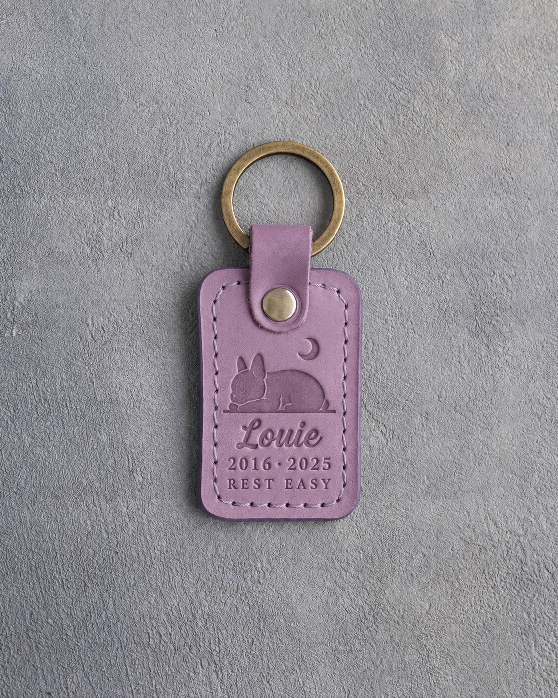 Lilac leather keychain debossed with a French bulldog curled asleep under a crescent moon above Louie, 2016 · 2025 and REST EASY, on gray concrete
