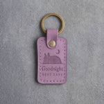 Lilac leather keychain debossed with a French bulldog curled asleep under a crescent moon above Goodnight and REST EASY, on gray concrete