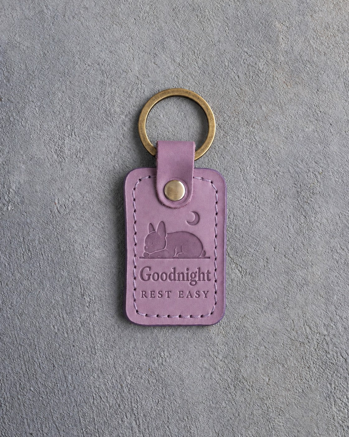 Lilac leather keychain debossed with a French bulldog curled asleep under a crescent moon above Goodnight and REST EASY, on gray concrete
