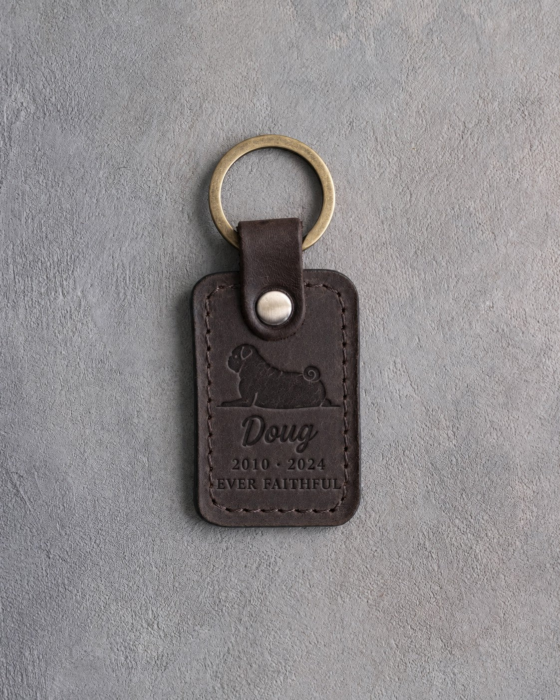 Dark espresso leather keychain debossed with a pug lying head up above Doug, 2010 · 2024 and EVER FAITHFUL, on gray concrete