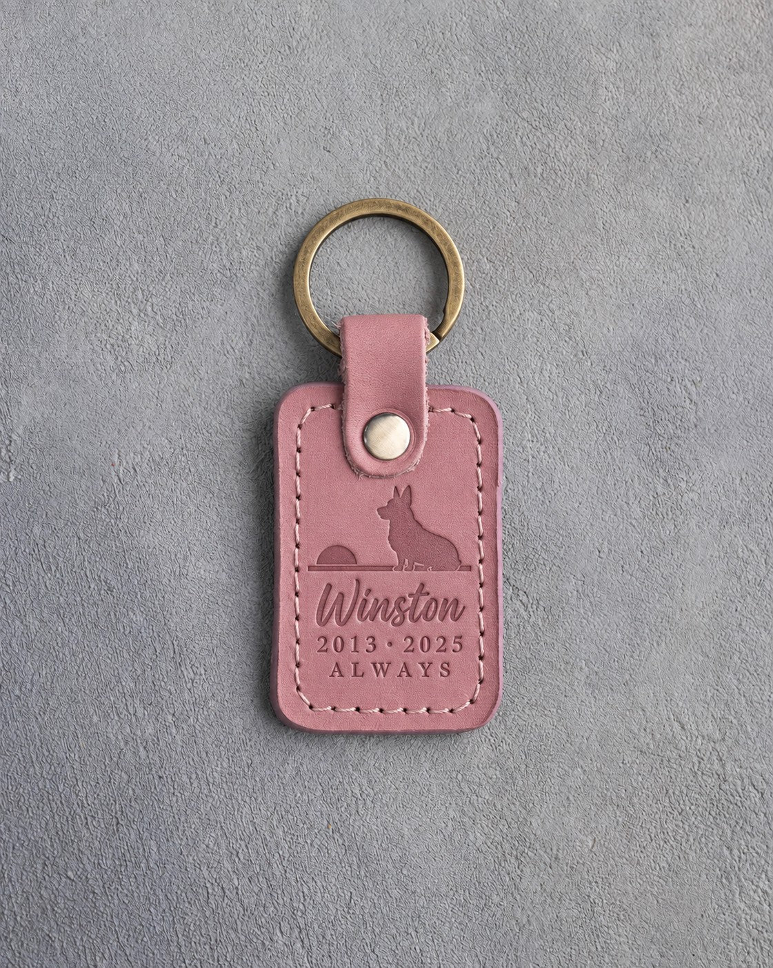 Parisian Blossom pink leather keychain debossed with a corgi seated on a horizon facing a low sun above Winston, 2013 · 2025 and ALWAYS, on gray concrete
