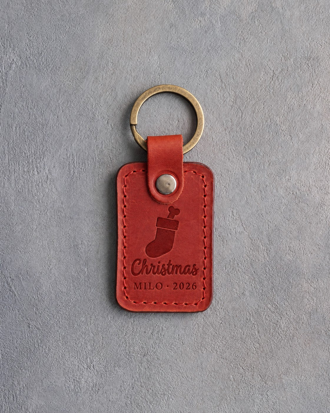 Poppy red leather keychain debossed with a Christmas stocking holding a dog bone above Christmas and MILO · 2026, on gray concrete