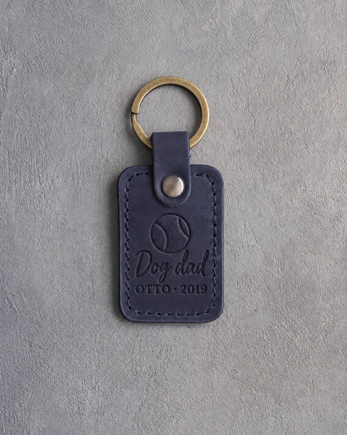 Blueberry navy leather keychain debossed with a tennis ball above Dog dad and OTTO · 2019, on gray concrete