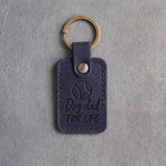 Blueberry navy leather keychain debossed with a tennis ball above Dog dad and FOR LIFE, on gray concrete