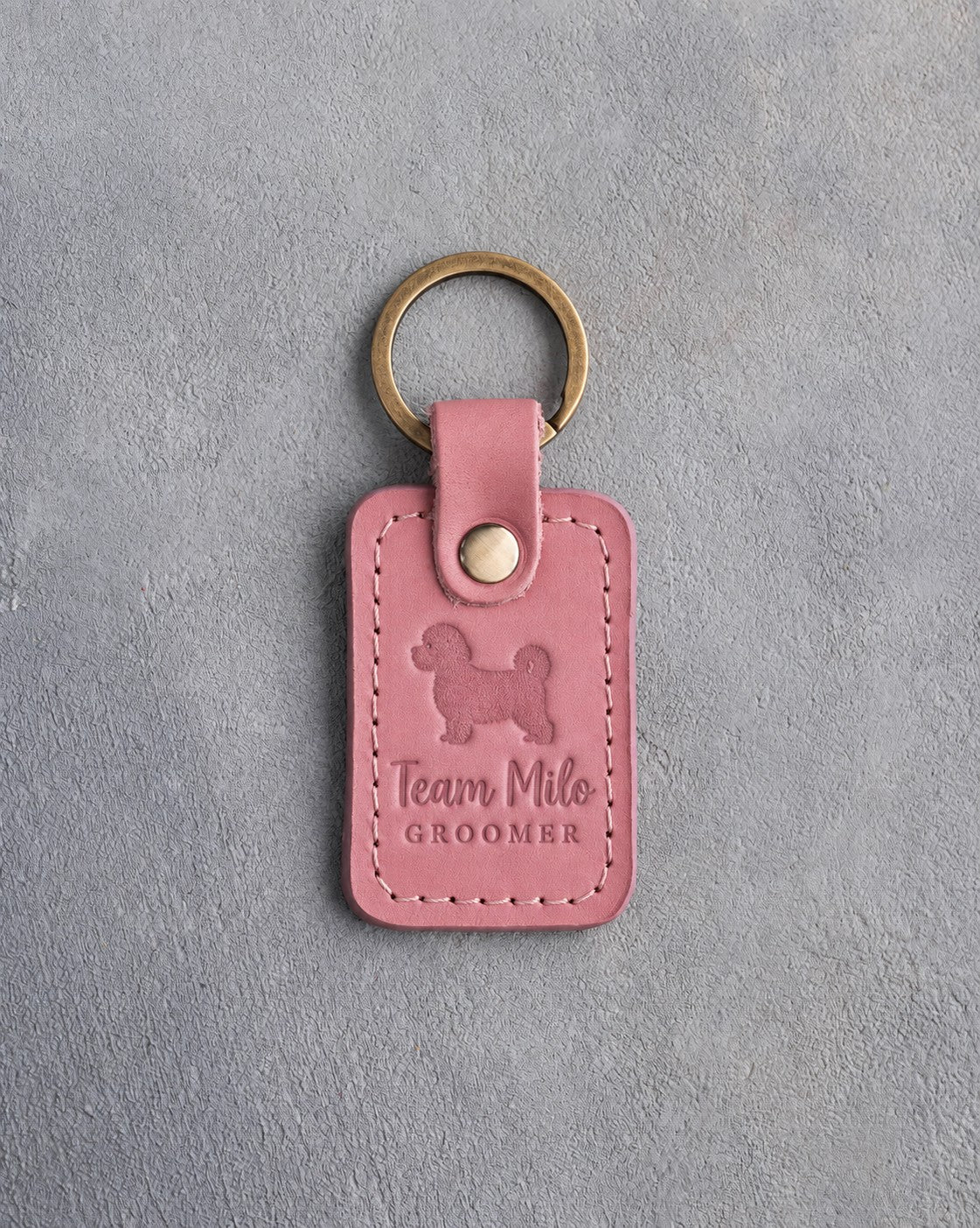 Parisian Blossom pink leather keychain debossed with a maltipoo above Team Milo and GROOMER, on gray concrete