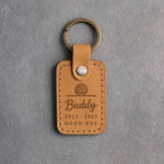 Indian Summer tan leather keychain debossed with a ball resting on the floor above Buddy, 2012 · 2025 and GOOD BOY, on gray concrete