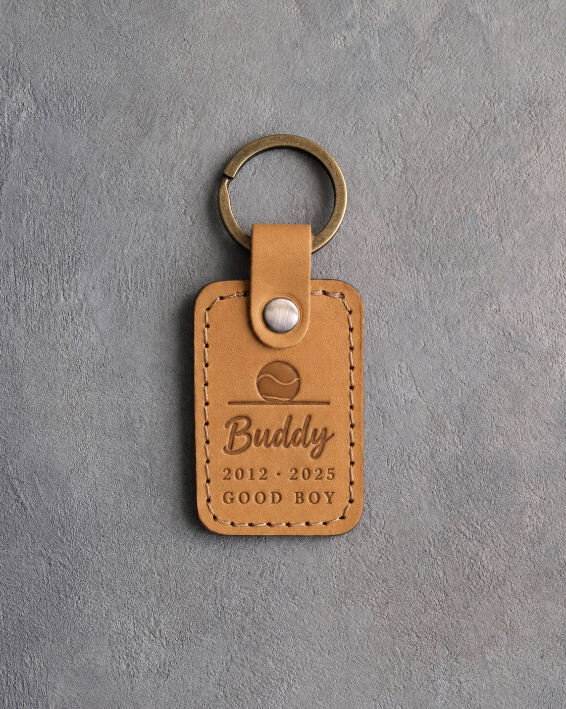 Indian Summer tan leather keychain debossed with a ball resting on the floor above Buddy, 2012 · 2025 and GOOD BOY, on gray concrete