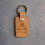 Indian Summer tan leather keychain debossed with a ball resting on the floor above Until we play and AGAIN, on gray concrete