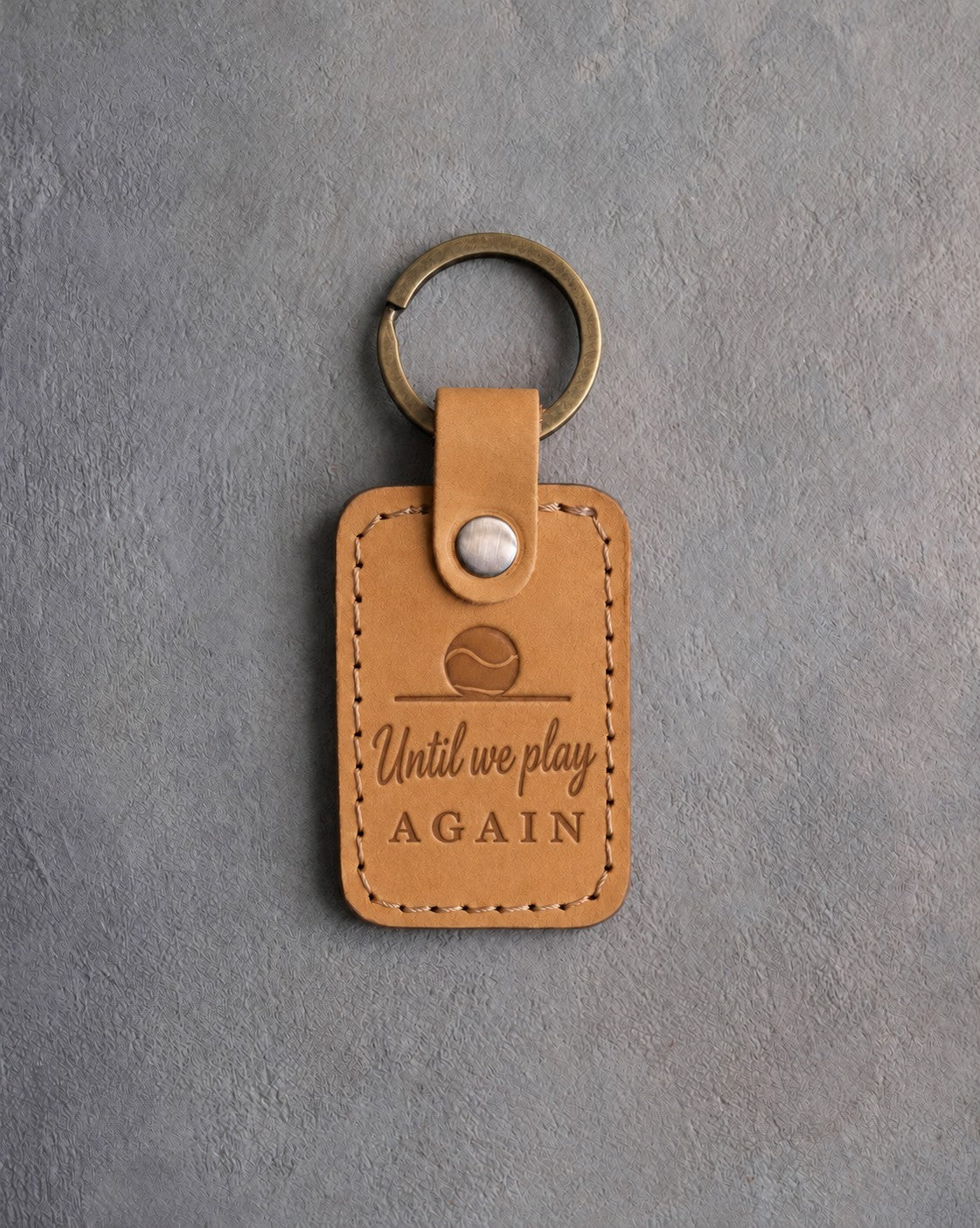 Indian Summer tan leather keychain debossed with a ball resting on the floor above Until we play and AGAIN, on gray concrete