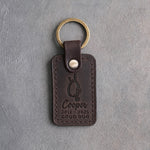 Dark espresso leather keychain debossed with an empty dog collar on a hook above Cooper, 2012 · 2025 and GOOD DOG, on gray concrete