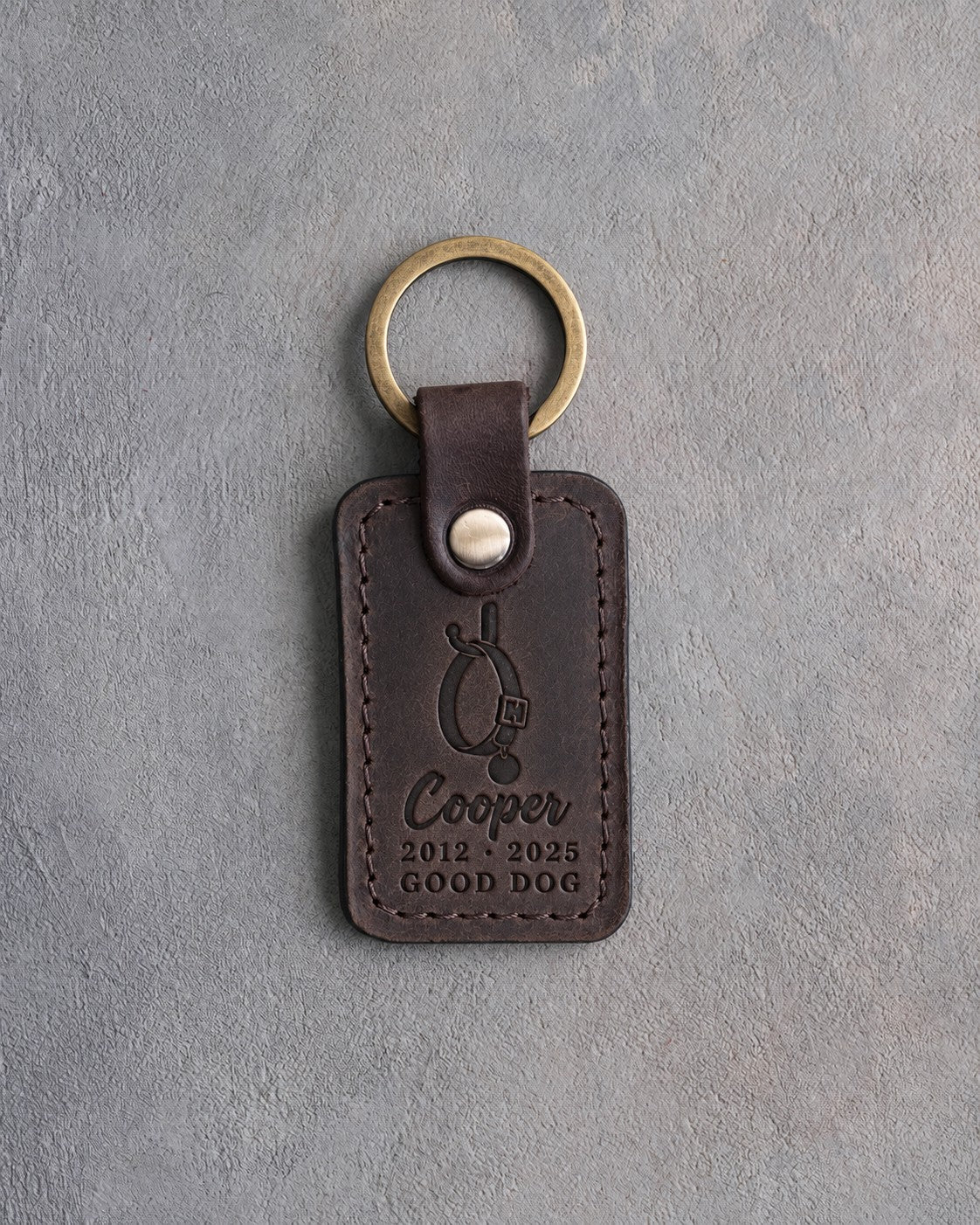 Dark espresso leather keychain debossed with an empty dog collar on a hook above Cooper, 2012 · 2025 and GOOD DOG, on gray concrete