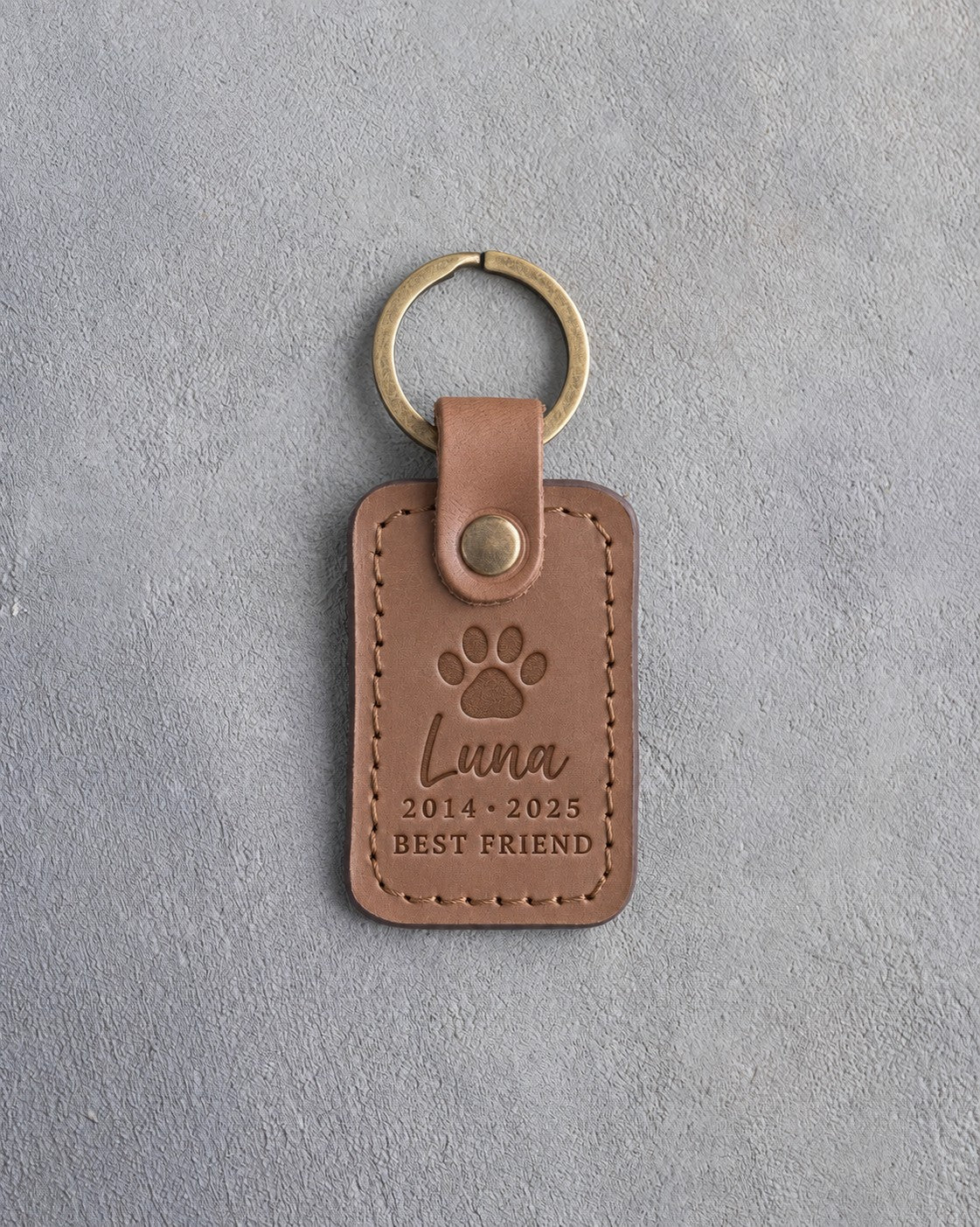 Caramel leather keychain debossed with a single paw print above Luna, 2014 · 2025 and BEST FRIEND, on gray concrete