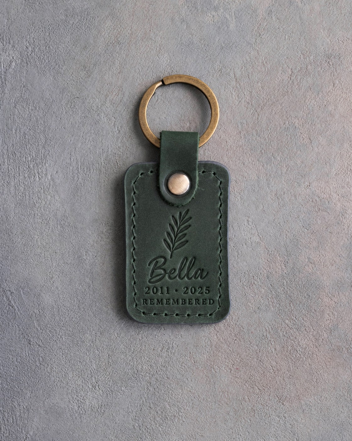 Forest green leather keychain debossed with a sprig of rosemary above Bella, 2011 · 2025 and REMEMBERED, on gray concrete