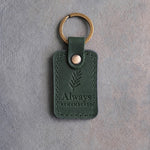 Forest green leather keychain debossed with a sprig of rosemary above Always and REMEMBERED, on gray concrete