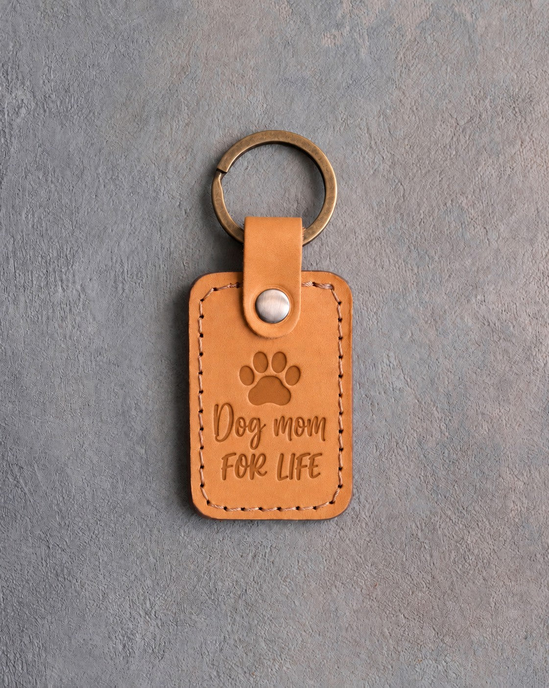 Indian Summer tan leather keychain debossed with a paw print above Dog mom and FOR LIFE, on gray concrete