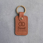 Caramel leather keychain debossed with two interlocked wedding rings above the name Biscuit and DOG OF HONOR, on gray concrete