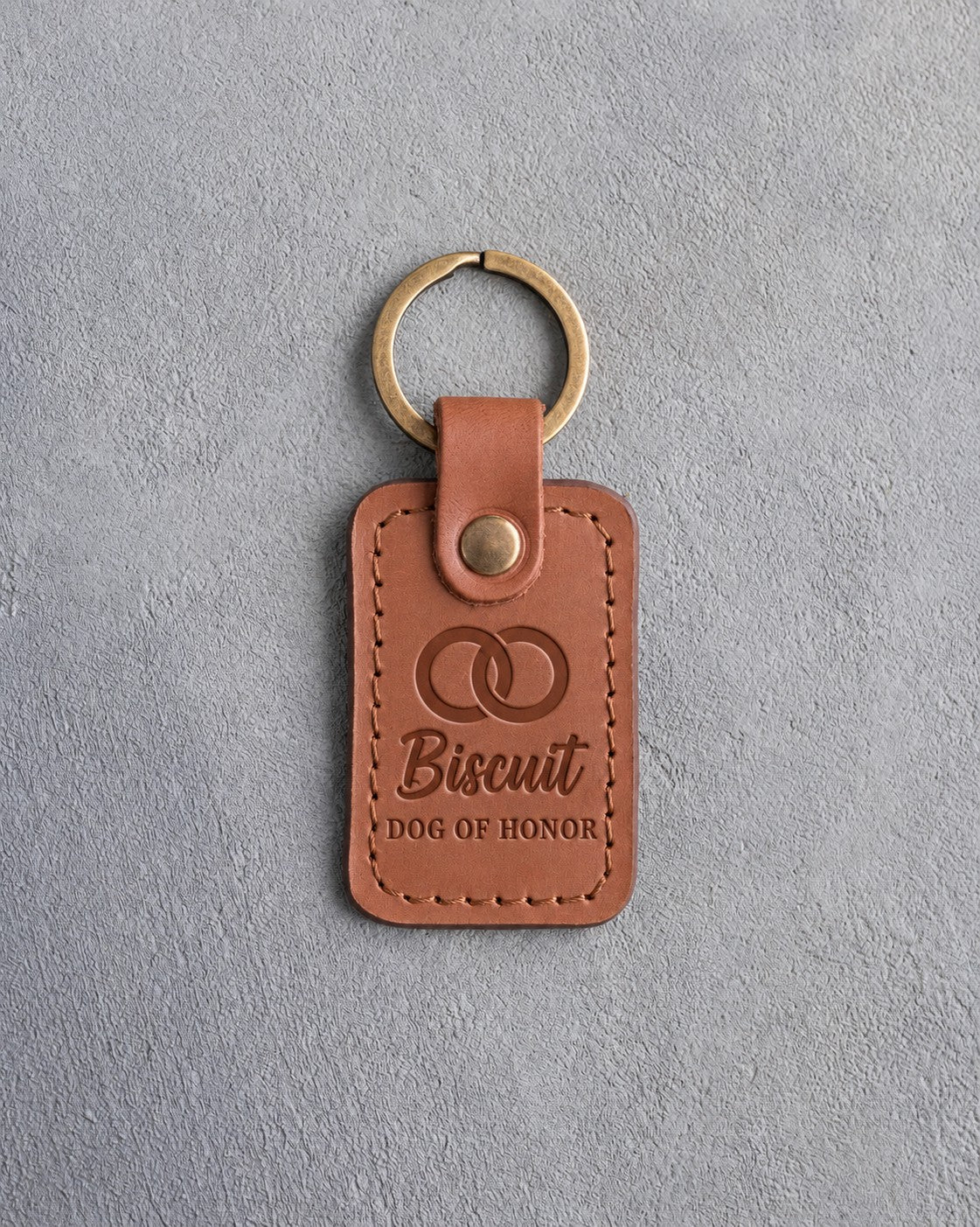 Caramel leather keychain debossed with two interlocked wedding rings above the name Biscuit and DOG OF HONOR, on gray concrete