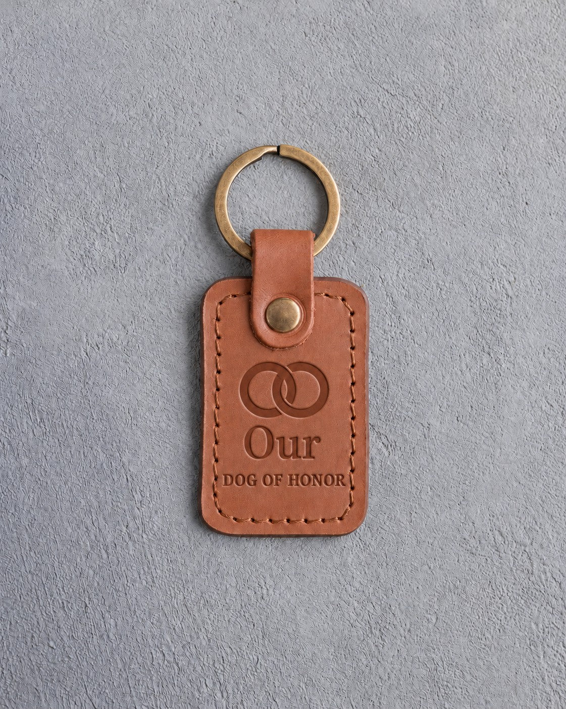 Caramel leather keychain debossed with two interlocked wedding rings above Our and DOG OF HONOR, on gray concrete