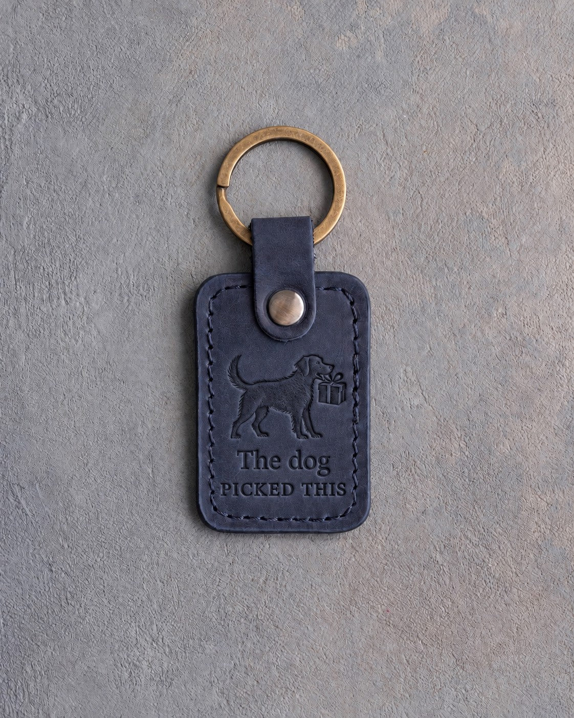 Blueberry navy leather keychain debossed with a dog carrying a wrapped gift above The dog and PICKED THIS, on gray concrete