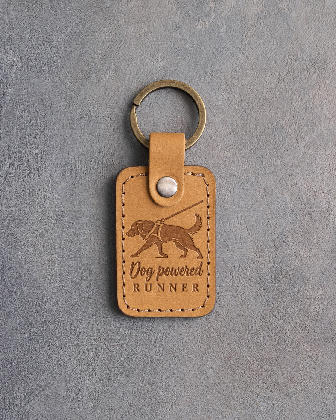 Indian Summer tan leather keychain debossed with a dog pulling on a harness line and the words Dog powered RUNNER, on gray concrete