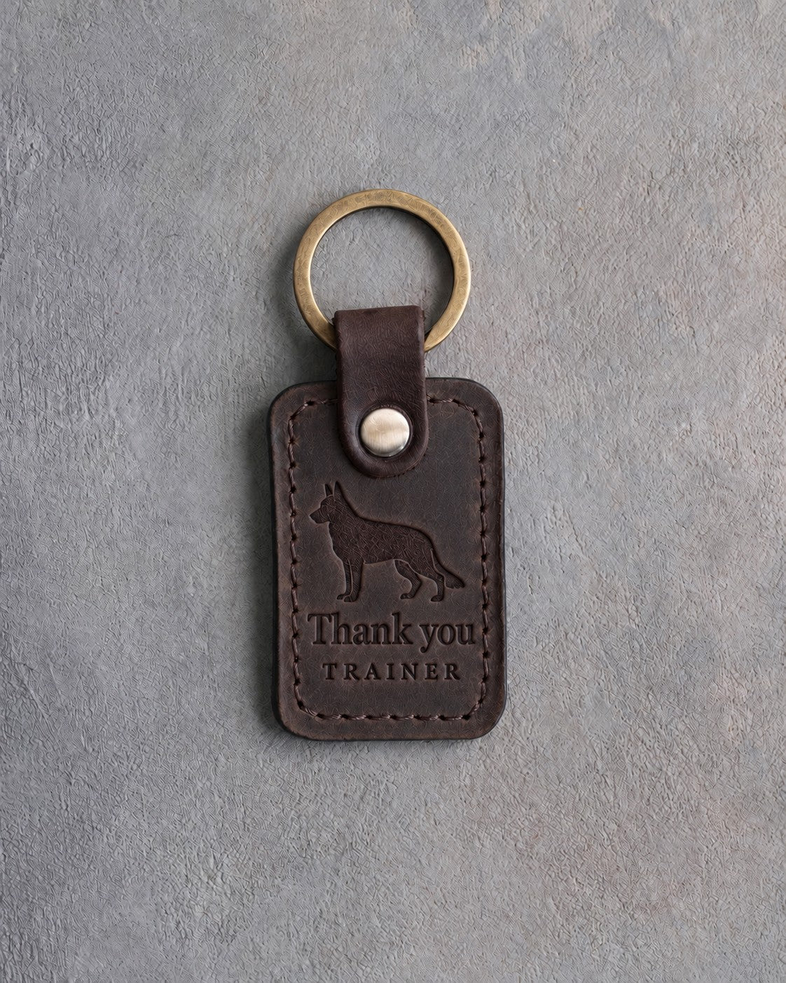 Dark espresso leather keychain debossed with a German shepherd above Thank you and TRAINER, on gray concrete