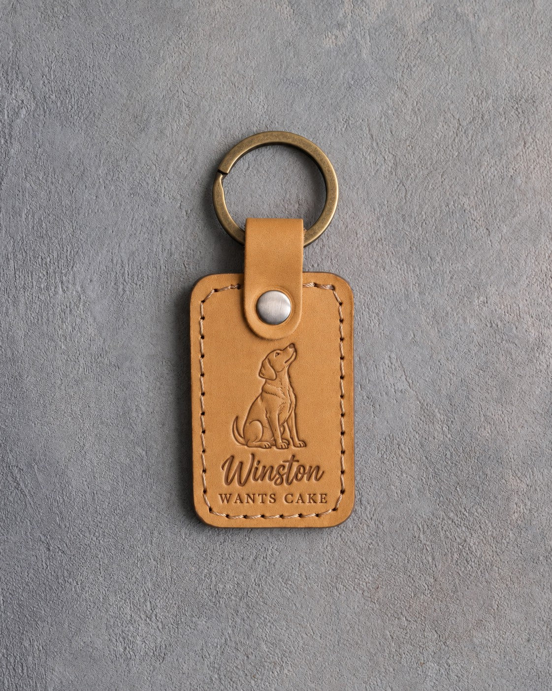 Indian Summer tan leather keychain debossed with a begging dog above Winston and WANTS CAKE, on gray concrete