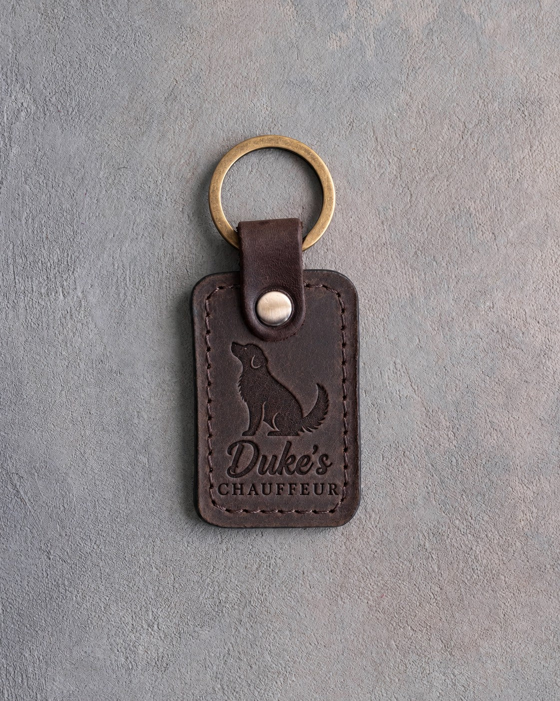 Dark espresso leather keychain debossed with a sitting dog looking up above Duke's and CHAUFFEUR, on gray concrete