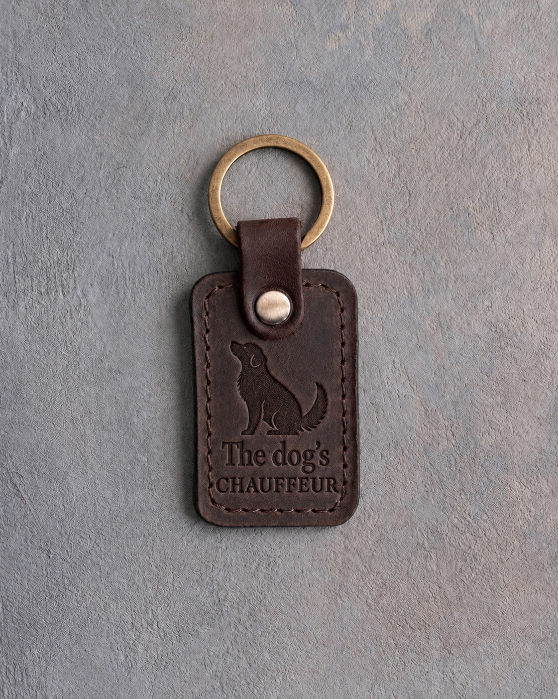 Dark espresso leather keychain debossed with a sitting dog looking up above The dog's and CHAUFFEUR, on gray concrete