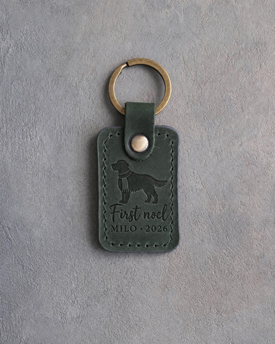 Forest green leather keychain debossed with a golden retriever in a knitted scarf above First noel and MILO · 2026, on gray concrete