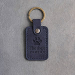 Blueberry navy leather keychain debossed with a paw print above The dog's and PERSON, on gray concrete