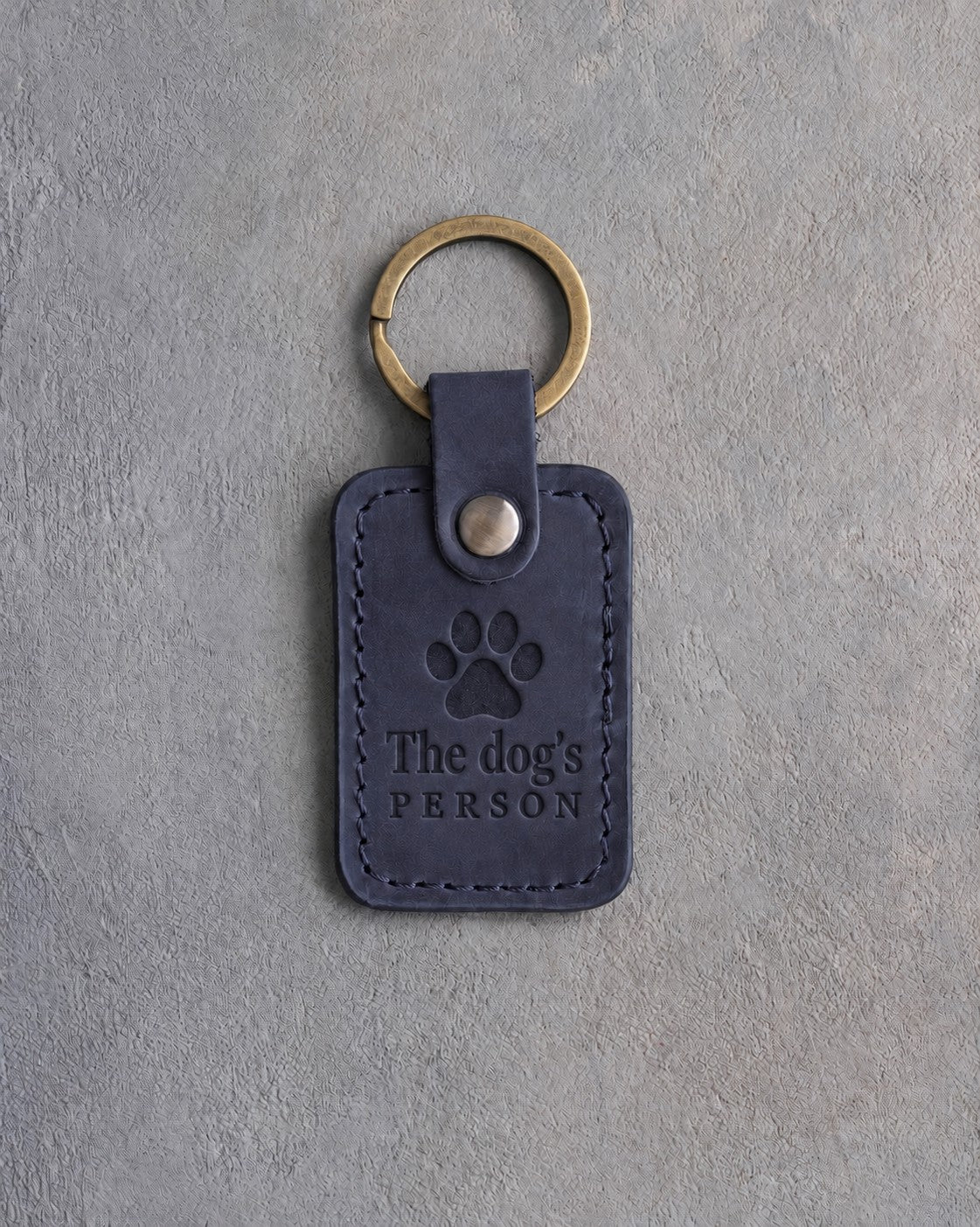 Blueberry navy leather keychain debossed with a paw print above The dog's and PERSON, on gray concrete