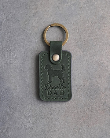 Forest green leather keychain debossed with a goldendoodle silhouette and Doodle DAD, on gray concrete